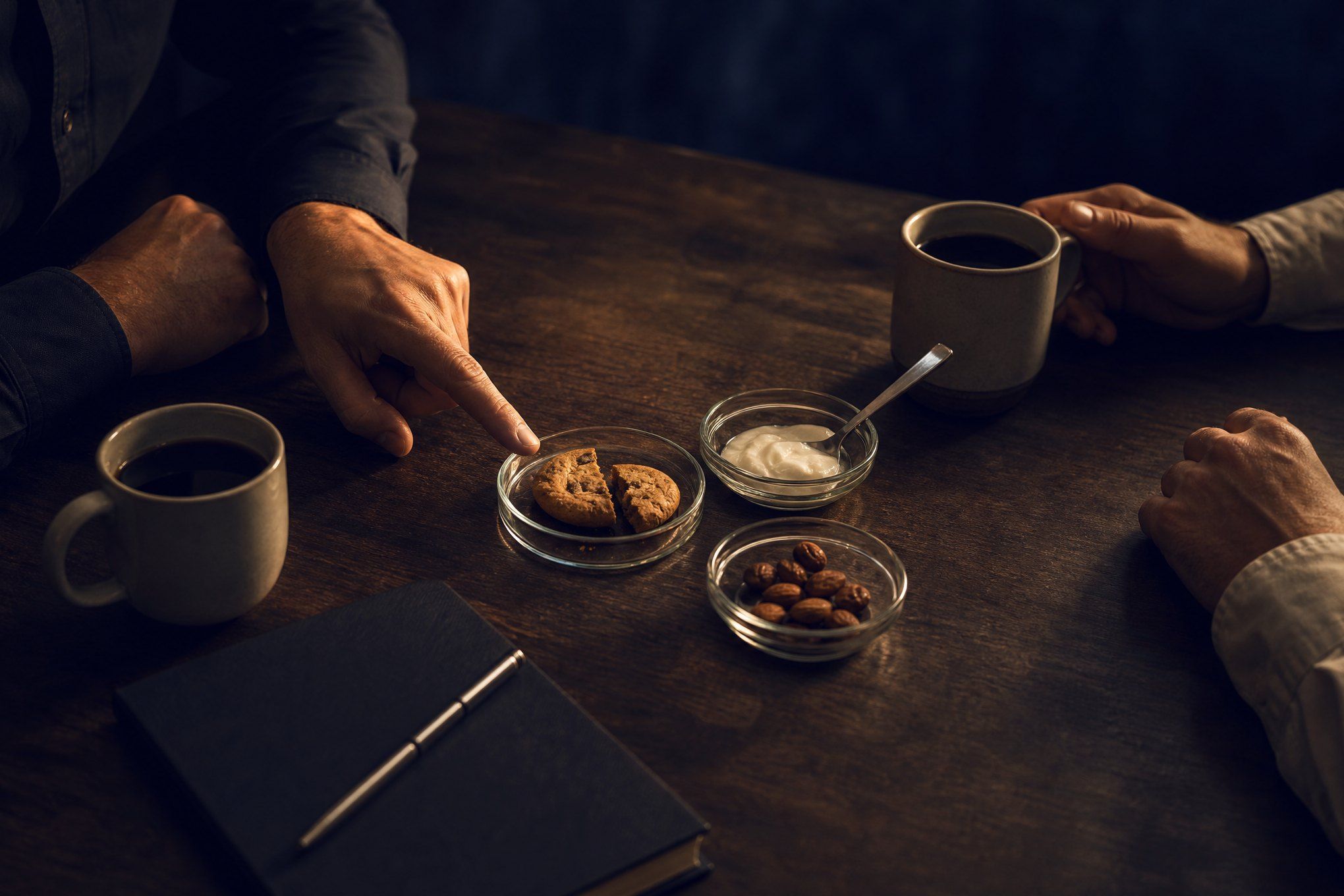 Generated image: Hands around a table with coffee cups, a closed notebook and small dishes of food samples; one hand points at a broken cookie.
